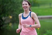 Emily, a woman in her twenties with brown hair, running towards the camera while smiling. She is wearing a salmon pink running vest and black shorts. Immediately behind her is a lake, with a grassy embankment and green trees in the background.
